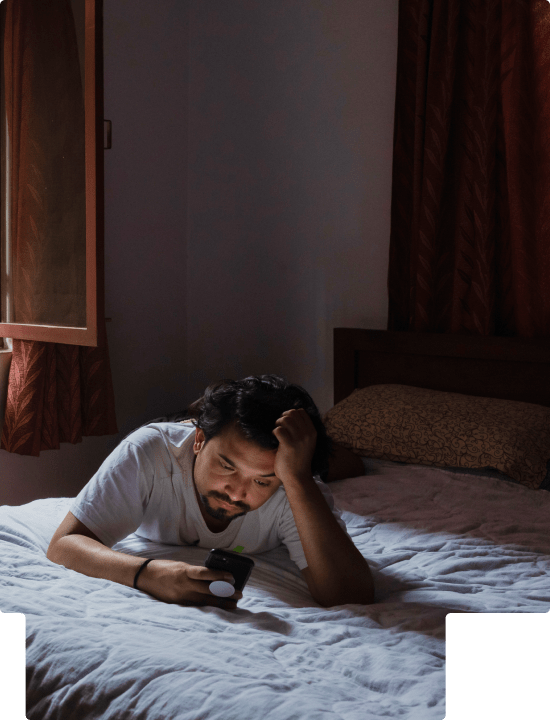 A man laying on his stomach on his bed holding his head with one hand and looking at his phone